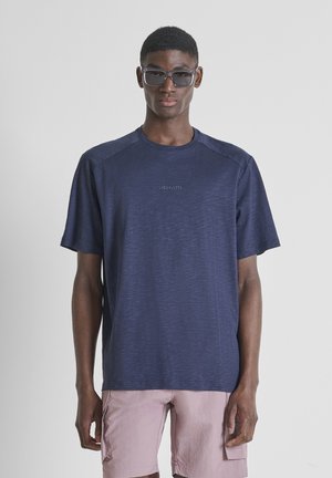 Čovjek u tamnoplavoj Morato majici, svijetloružičastim cargo kratkim hlačama i tamnim pravokutnim sunčanim naočalama stoji ispred sive pozadine.