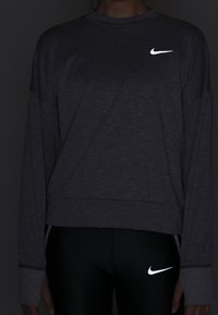 Felpa Nike grigia a maniche lunghe con logo riflettente sul petto, abbinata a leggings neri con un piccolo logo riflettente.
