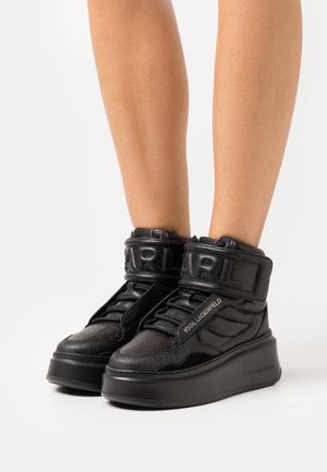 Zapatillas de plataforma negras de caña alta con correas acolchadas en el tobillo con el relieve de las marcas "KARL" y "KARL LAGERFELD" en los laterales, usadas sobre piernas desnudas.
