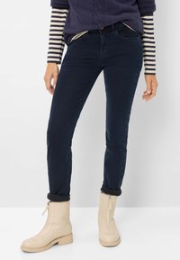 Slim-fit mørkeblå jeans med en glat tekstur, parret med cremede ankelstøvler med en sidezipper og blokhæl. Diskret opkrøllede detaljer ved kanten.