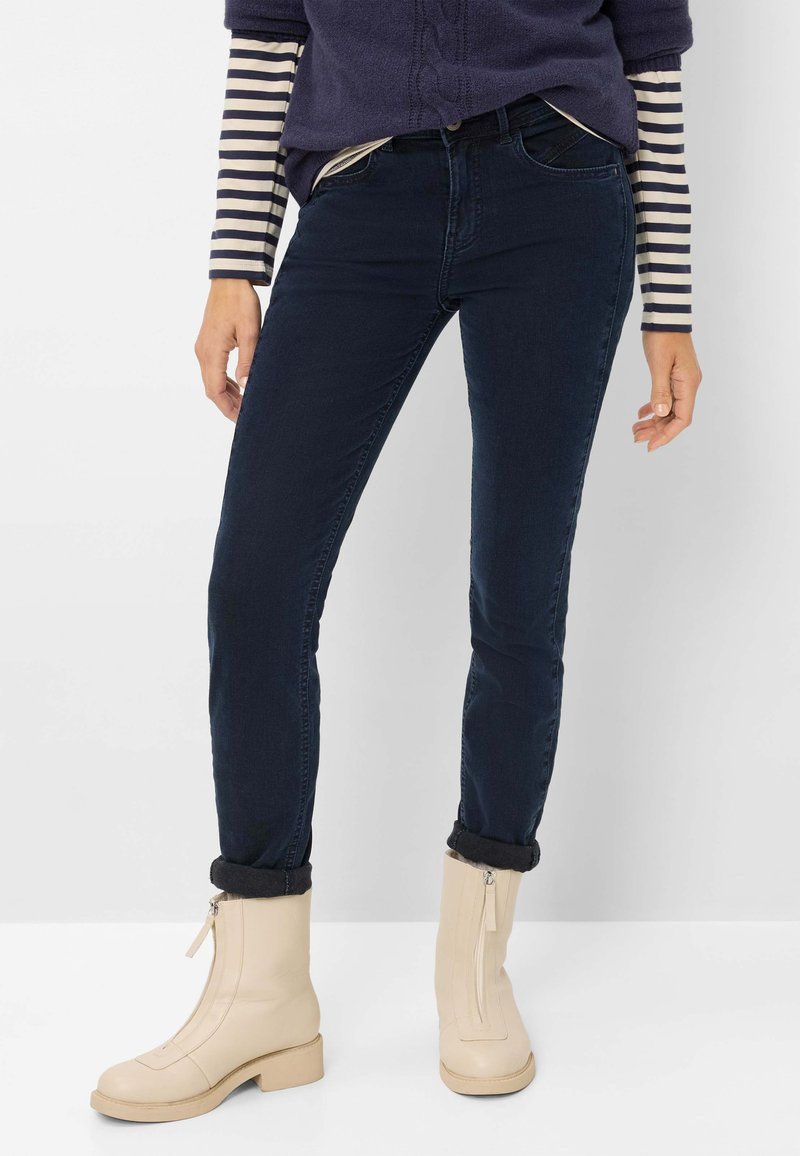 Slim-fit mørkeblå jeans med en glat tekstur, parret med cremede ankelstøvler med en sidezipper og blokhæl. Diskret opkrøllede detaljer ved kanten.