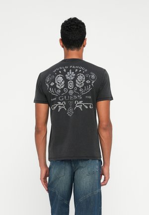 Hombre con cabello corto y rizado de pie de espaldas, vistiendo una camiseta gráfica negra con diseño floral y texto, y pantalones vaqueros azules.