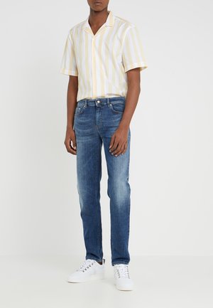 Homme portant une chemise à manches courtes rayée verticale beige et blanche, un jean slim bleu moyen et des baskets blanches sur un fond uni.