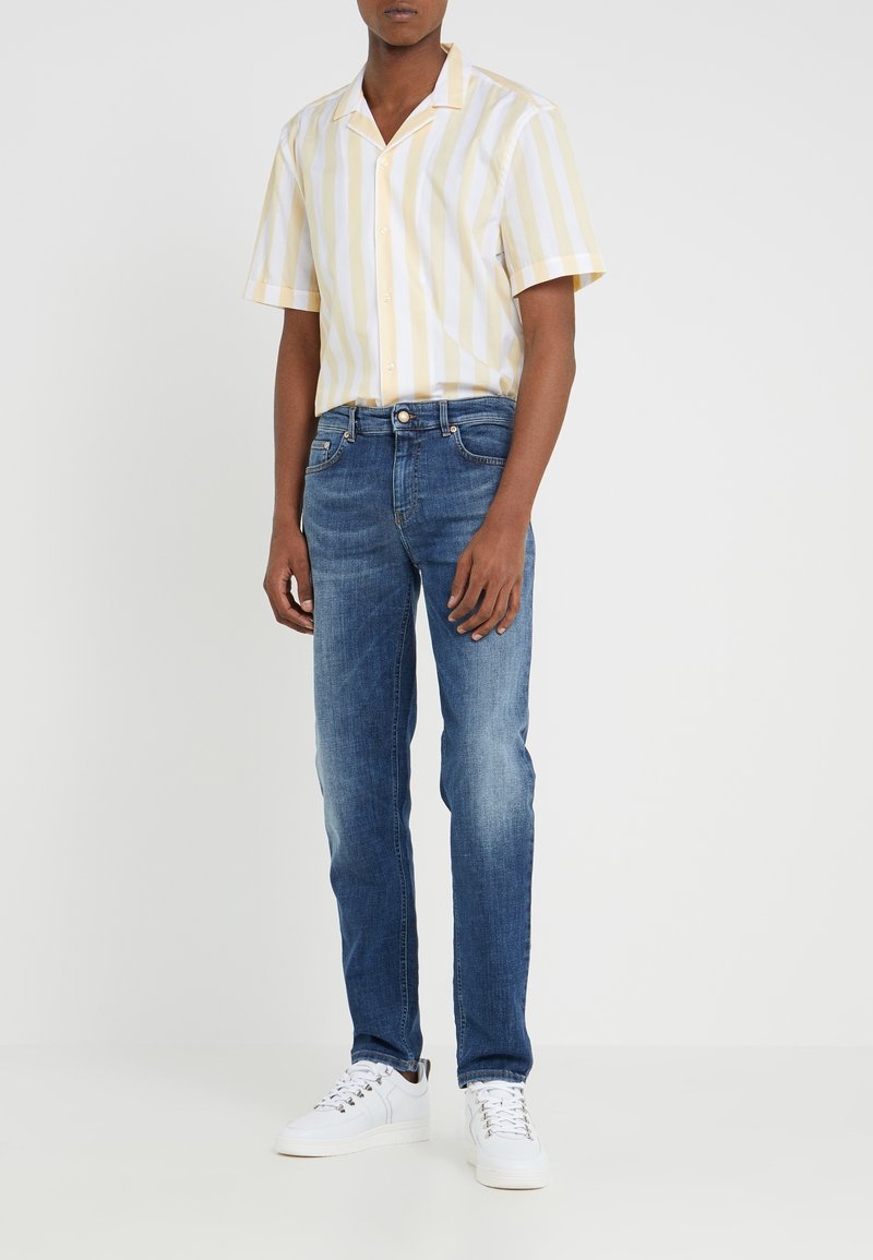 Homme portant une chemise à manches courtes rayée verticale beige et blanche, un jean slim bleu moyen et des baskets blanches sur un fond uni.