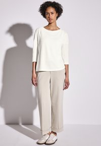 Donna con capelli ricci che indossa una maglietta bianca a tre quarti, pantaloni beige a gamba larga e sneakers beige e bianche, in piedi contro un muro bianco.