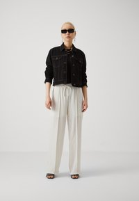 Veste cropped en denim noir avec des boutons, associée à un pantalon blanc à cordon de serrage. Des lunettes de soleil noires et des sandales plates complètent le look.