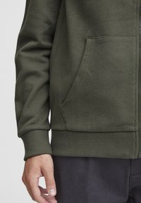 Hoodie zippé vert olive en tissu doux et texturé. Équipé d'une poche kangourou et de poignets et d'un ourlet côtelés pour un ajustement confortable.