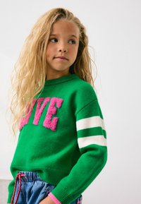 Green knitted sweater featuring "LOVE" in pink plush letters, white and green striped sleeves, and a round neckline.