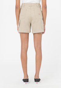 Pantaloni cargo beige con vita alta, tasche laterali e posteriori, e una texture di cotone liscia. Indossati con scarpe nere senza lacci.