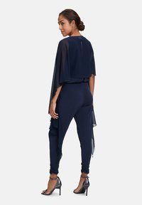 Marineblauer Overall mit geschichteter Chiffon-Cape. Mit schmalen Beinen, seitlichen Rüschen und eleganten High Heels. Glatte Textur und elegante Silhouette.