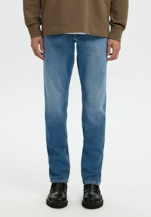 Personne portant un jean bleu délavé à jambes droites, un haut marron à manches longues et des chaussures en cuir noir, debout devant un fond uni.