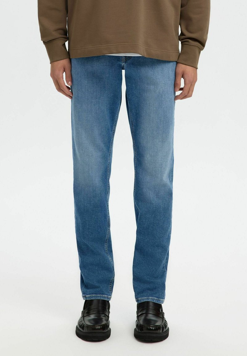 Personne portant un jean bleu délavé à jambes droites, un haut marron à manches longues et des chaussures en cuir noir, debout devant un fond uni.