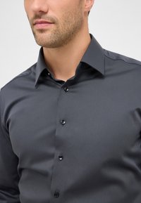 Camicia button-up grigia scura realizzata in tessuto liscio con colletto a punta e cinque bottoni neri. Presenta una trama sottile e un design aderente.