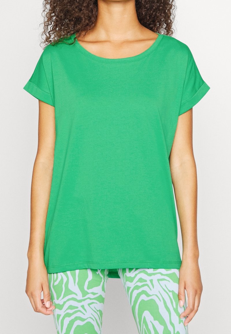 Femme portant un t-shirt vert vif à manches courtes et un pantalon à motifs vert clair et blanc, debout les bras détendus le long du corps.