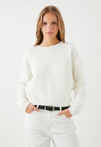 Weißer gerippter Pullover mit Rundhalsausschnitt, lockerer Passform und langen Ärmeln, kombiniert mit hochgeschnittenen weißen Jeans und einem schwarzen Gürtel.