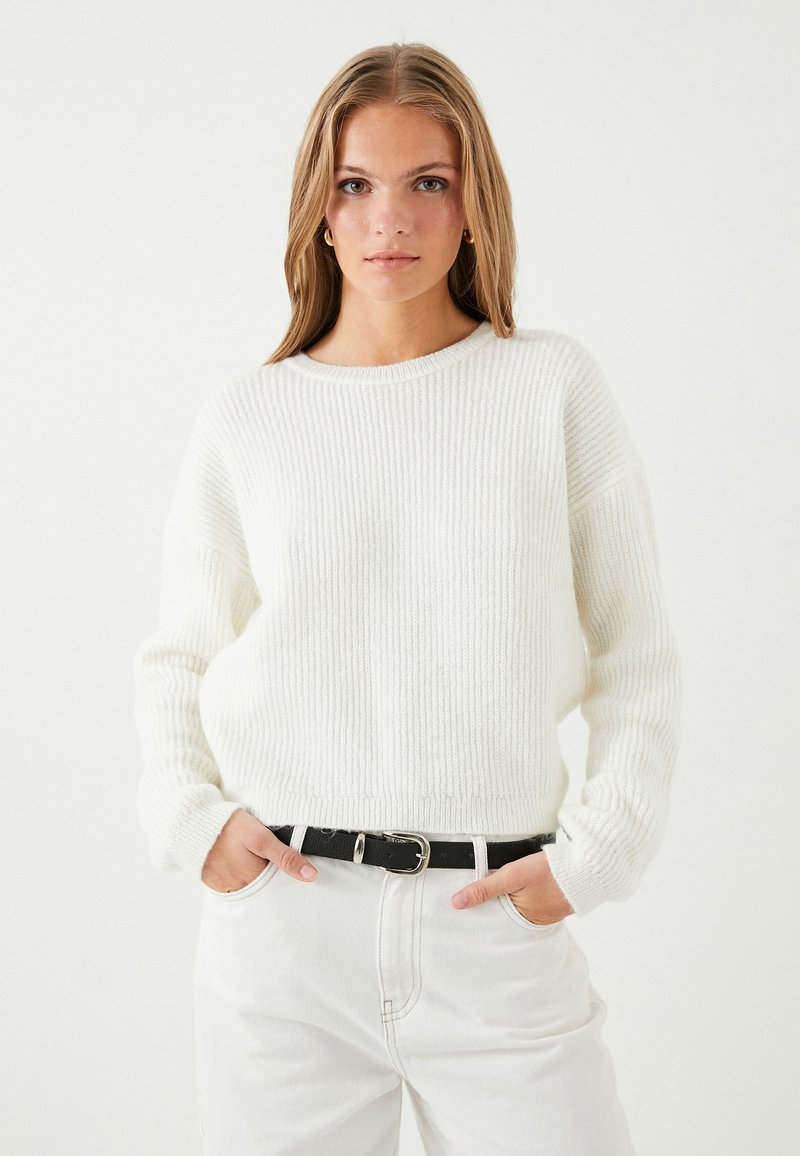 Weißer gerippter Pullover mit Rundhalsausschnitt, lockerer Passform und langen Ärmeln, kombiniert mit hochgeschnittenen weißen Jeans und einem schwarzen Gürtel.