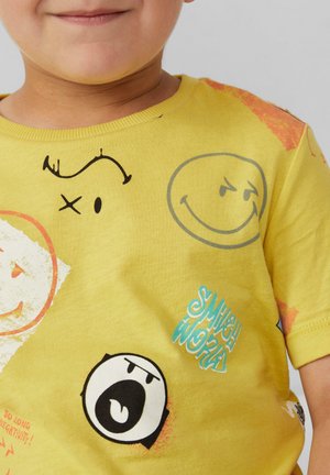 Geel katoenen T-shirt met verschillende bedrukte glimlachende gezichten en uitdrukkingen, met blauwe en zwarte tekstaccenten op de voorkant. Korte mouwen.