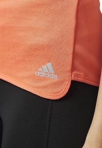 Lekki, teksturowany pomarańczowy top sportowy z podwiniętym dołem, z metalicznym logo Adidas na dolnym rogu oraz czarne legginsy.