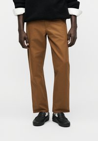 Άνδρας που φοράει καφέ παντελόνια, μαύρα μοκασίνια, λευκές κάλτσες, μαύρο πουλόβερ με λευκά μανίκια πουκάμισου, στέκεται μπροστά σε απλό φόντο.