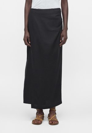 Άτομο που φορά μακριά μαύρη φούστα, λευκό τοπ και μπεζ φλατ σανδάλια με λουράκια, στέκεται πάνω σε ένα απλό φωτεινό φόντο.