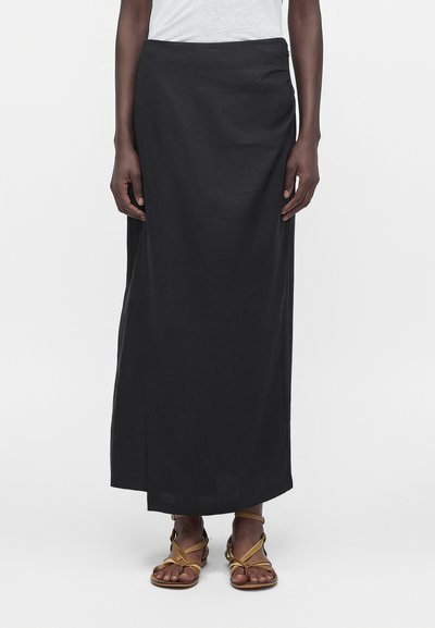 Person iført en lang sort nederdel, hvid top og beige flade sandaler med remme, stående mod en ensfarvet lys baggrund.