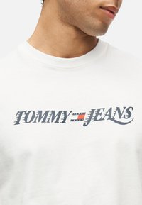 Κοντινό πλάνο ατόμου που φοράει λευκό πουκάμισο με το λογότυπο "Tommy Jeans" σε στιλιζαρισμένη μαύρη γραμματοσειρά και ένα μικρό έμβλημα με κόκκινη, λευκή και μπλε σημαία.