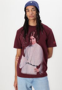 Maroon cotton t-shirt featuring a graphic print of a woman in a white outfit with long sleeves and hairstyle, complemented by light blue jeans.