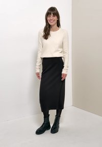 Femme souriante aux longs cheveux bruns portant un pull crème, une jupe midi noire, des collants noirs et des bottes noires à lacets, debout contre un mur uni.