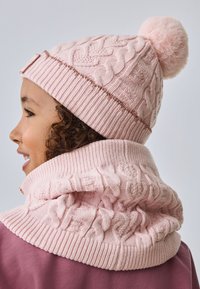 Svetlo roza kvačkana kapa z vzorcem vrvi, na vrhu okrašena s puhasto cofko. V kompletu z ujemajočim se puloverjem z visokim ovratnikom in rebrasto teksturo.