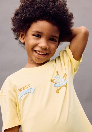 Enfant souriant aux cheveux bouclés portant un t-shirt jaune avec le texte "WEST County" et un badge en forme d'étoile de shérif, posant avec un bras derrière la tête.