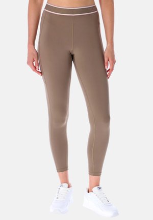Femme portant des leggings taille haute taupe avec une ceinture bordée de rose et des baskets blanches Reebok, debout devant un fond uni.