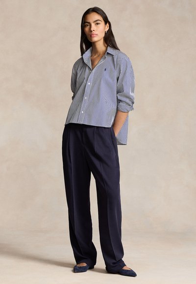 Žena stojaca s rukami v vreckách, oblečená v voľnej pruhovanej košeli, širokých námornícky modrých nohaviciach a námornícky modrých balerínach na neutrálnej pozadí.