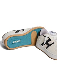 Witte sneaker met zwarte accenten, voorzien van een textuur, een rubberen zool met een blauwe binnenkant en logodetails aan de zijkant.