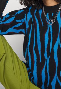 Un pull en tricot présentant des rayures de tigre bleues, noires et bourgogne. Le col rond est noir, associé à un pantalon vert sur mesure. Collier en chaîne métallique.