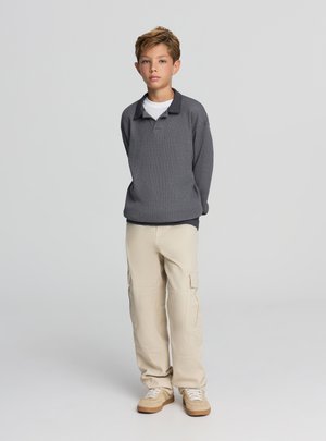 Polo grigia a maniche lunghe indossata sopra una t-shirt bianca, abbinata a pantaloni cargo beige e sneakers color cuoio. Stile casual, vestibilità rilassata.