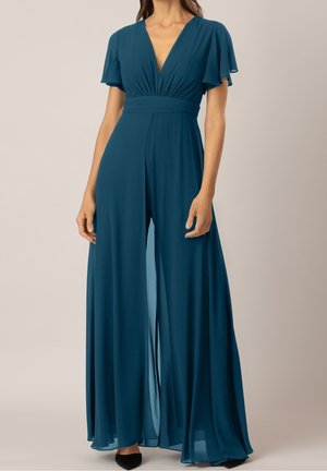 Combinaison pantalon large couleur sarcelle avec décolleté en V profond, manches papillon, taille cintrée et tissu fluide semi-transparent portée avec des chaussures noires à bout pointu.