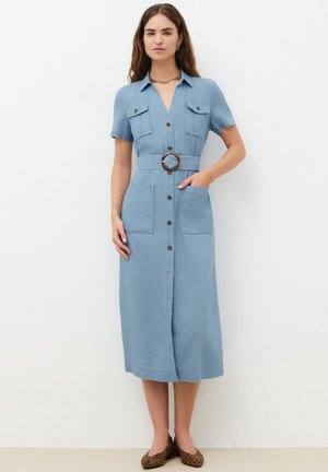Femme portant une robe midi en denim bleu clair, ceinturée et boutonnée, manches courtes, avec des ballerines à imprimé léopard, debout sur un fond blanc.