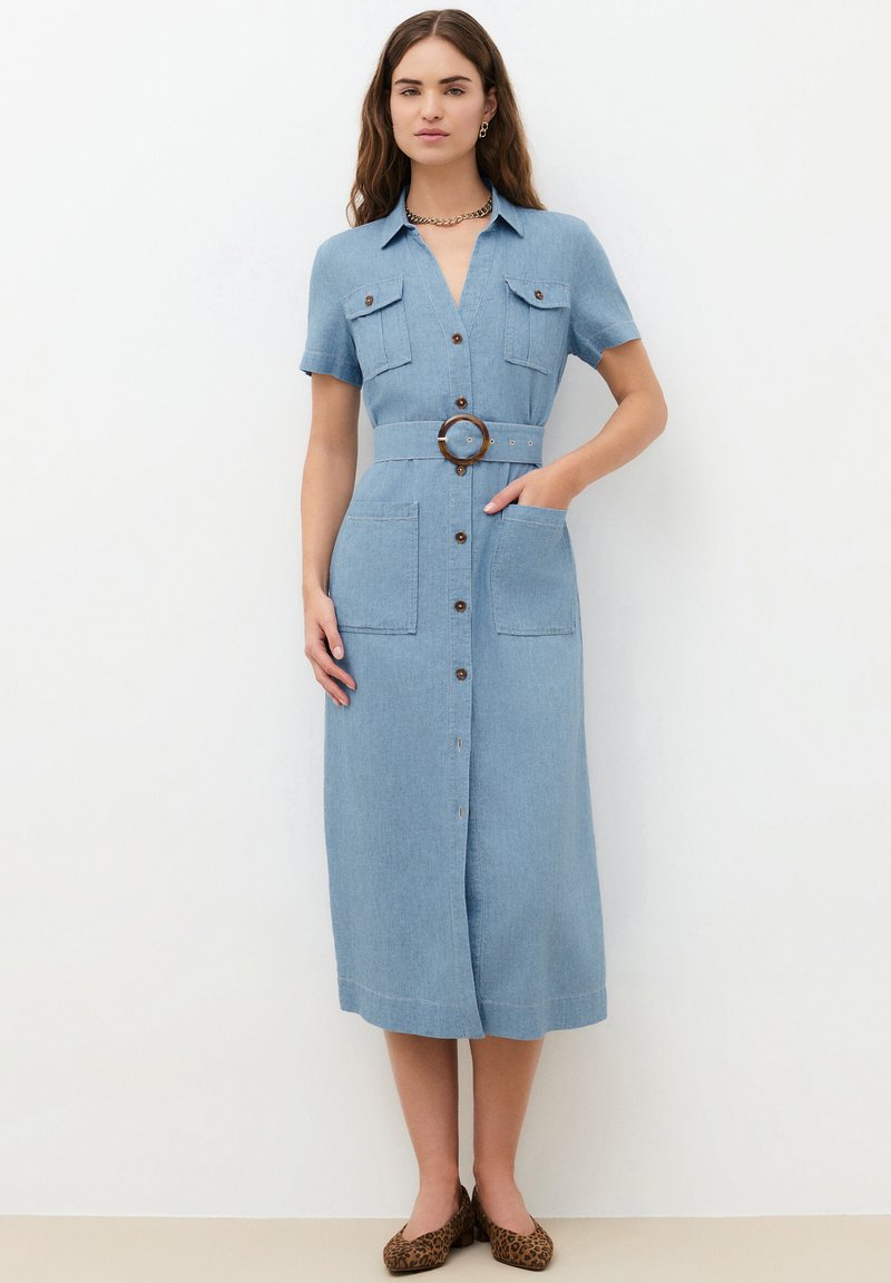 Femme portant une robe midi en denim bleu clair, ceinturée et boutonnée, manches courtes, avec des ballerines à imprimé léopard, debout sur un fond blanc.