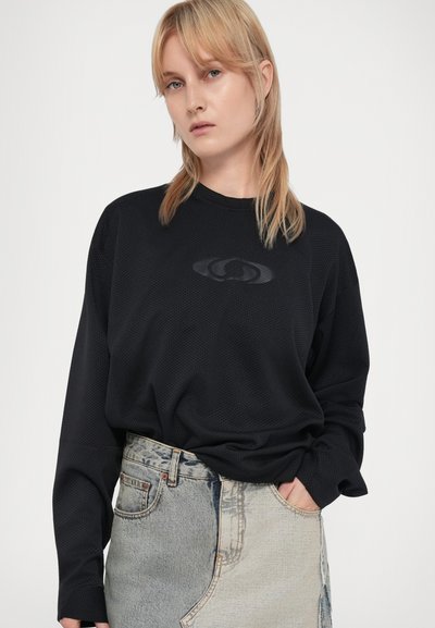 Zwarte langemouwen shirt van gestructureerde stof, met een subtiele logo in het midden, gecombineerd met een versleten lichtblauwe spijkerrok.