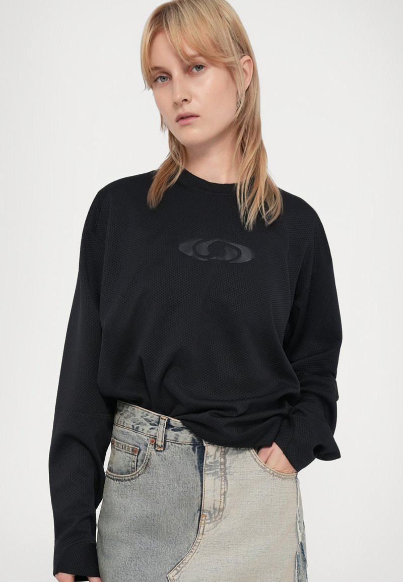 Sort langærmet skjorte lavet af tekstureret stof, med et diskret logo i midten, parret med en slidte lysvasket denimnederdel.
