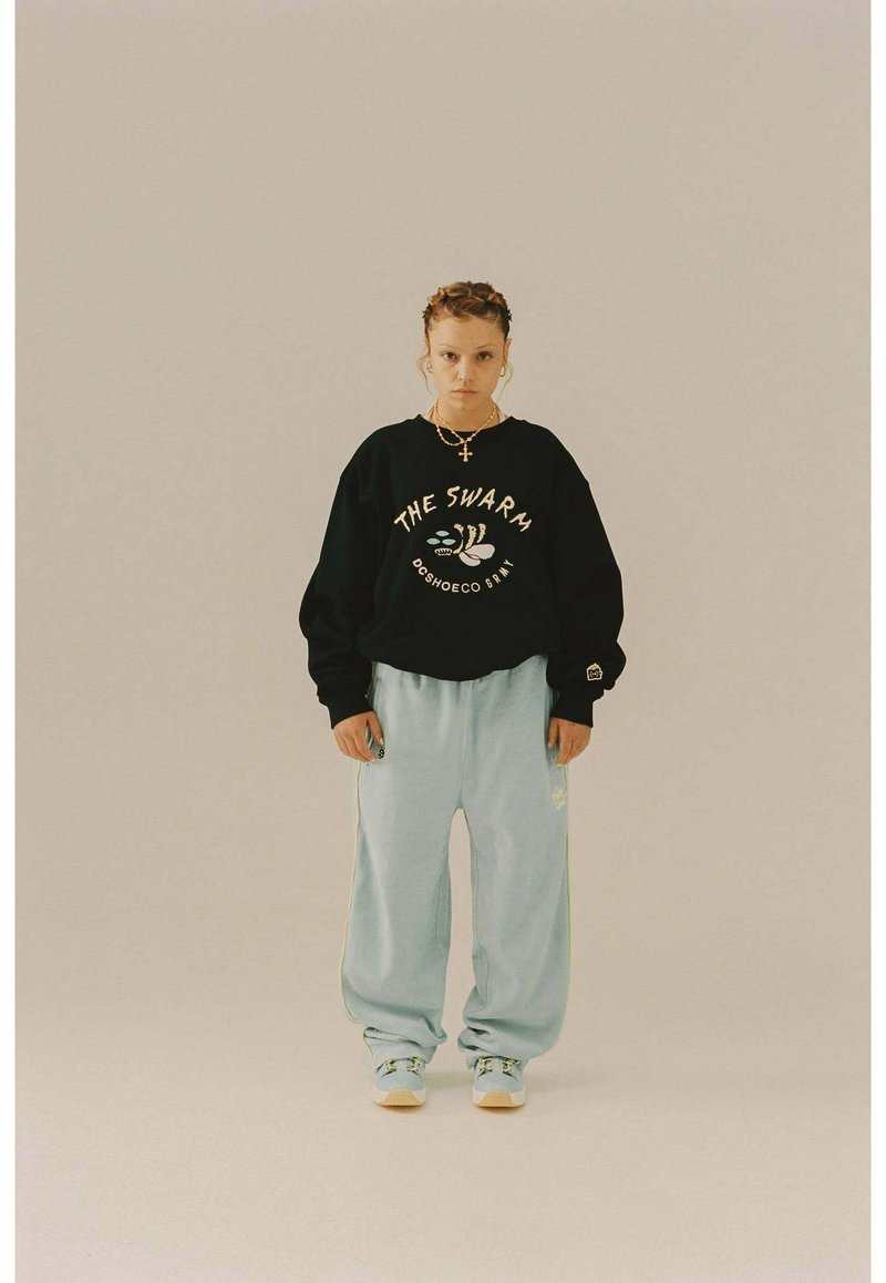 Sudadera negra con gráfico de "THE SWARM", combinada con pantalones deportivos sueltos de color azul claro. Presenta detalles dorados y una silueta relajada.