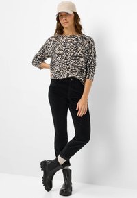 Pantalons en velours côtelé noir et un pull en imprimé animal beige. Le modèle porte une casquette beige et des bottines noires à semelle texturée.