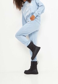 Stivali alla caviglia neri isolati con una finitura lucida, parte superiore testurizzata, una cerniera laterale e una suola spessa in gomma. Indossati con un abbigliamento da lounge di colore azzurro chiaro.
