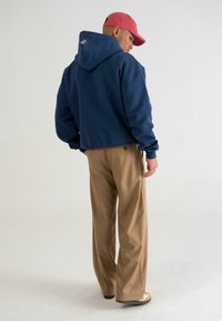 Felpa corta blu navy con tasca sul retro, abbinata a pantaloni beige a gamba larga, vista da dietro. Cappellino rosso che valorizza il look.