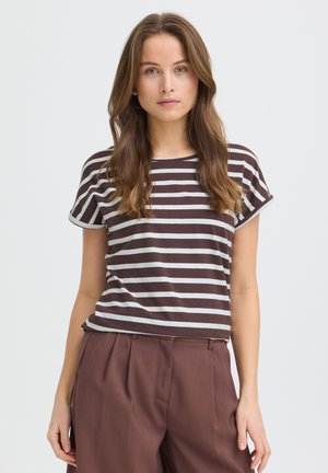 Mladá žena s dlhými hnedými vlasmi, oblečená v hnedo-bielom pruhovanom tričku s krátkym rukávom a hnedých vysokých nohaviciach, stojaca pred jednofarebným pozadím.