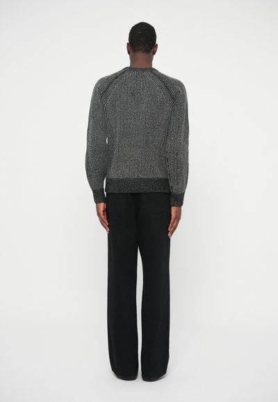 Υφασμάτινο, σκούρο γκρι πουλόβερ με ραφή, στρογγυλή λαιμόκοψη και εφαρμοστά μανίκια, συνδυασμένο με μαύρους παντελόνες σε φαρδιά γραμμή. Εμφανίζεται η πίσω όψη.