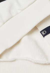 Tessuto misto cotone in off-white con colletto a coste e interno morbido e felpato. Presenta un'etichetta in tessuto blu con un emblema chiaro.
