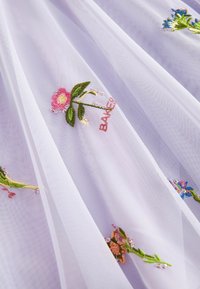 Weißer, transparenter Stoff mit floralem Stickmuster in Pink, Blau und Grün, feinen Perlenbesatz und dem Wort "BAKER" in Pink gestickt.
