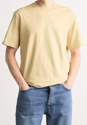 Osoba z rękami w kieszeniach, ubrana w jednolitą jasnżółtą koszulkę z krótkim rękawem i niebieskie dżinsy na neutralnym tle.