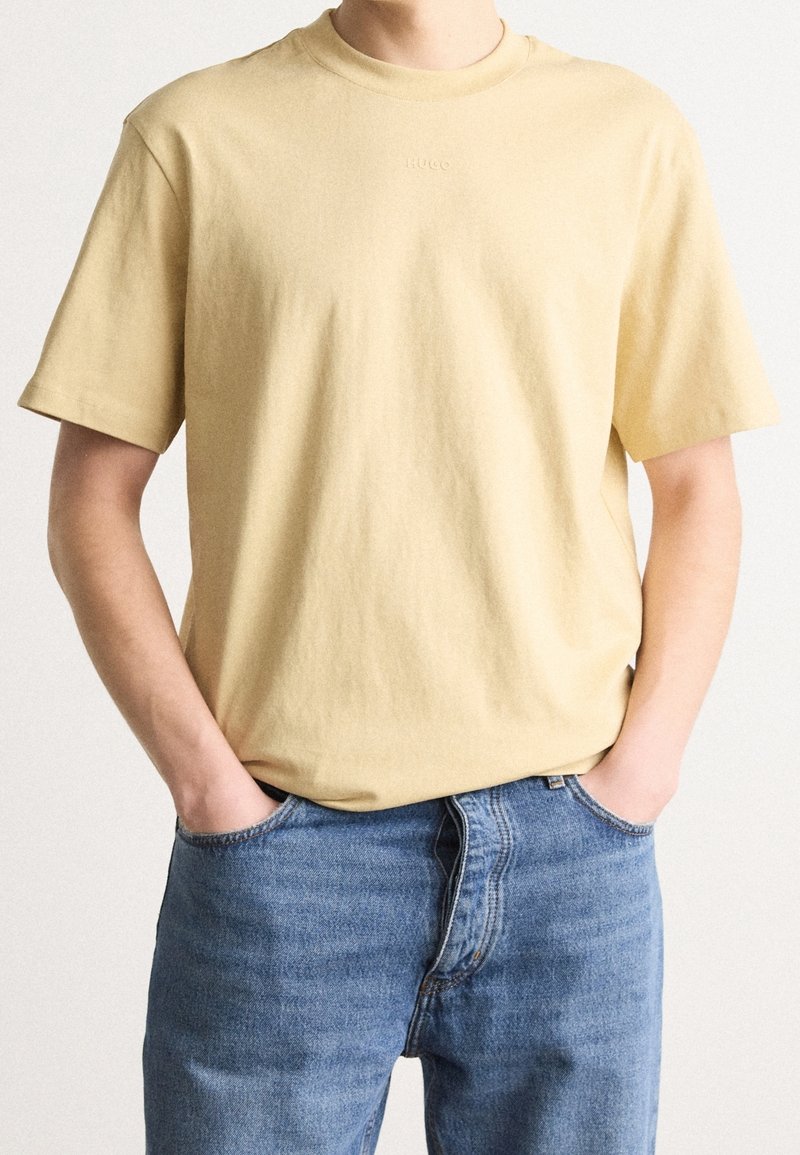 Personne avec les mains dans les poches portant un t-shirt à manches courtes jaune clair uni et un jean denim bleu sur un fond neutre.